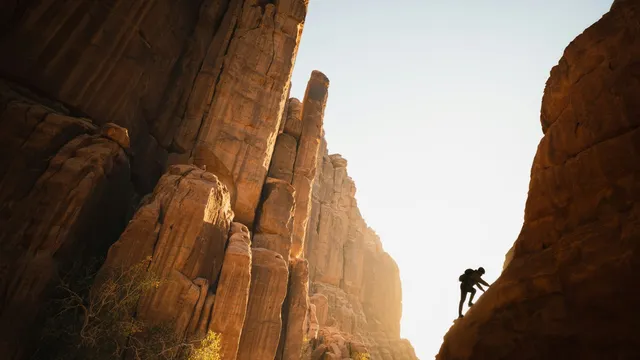 A lone climber silhouetted high on a sandstone cliff against a bright, hazy sky.