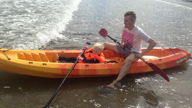 A man and a toddler in a life jacket sit in an orange kayak at the water's edge.