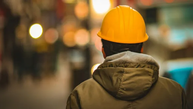 A man in a yellow hard hat and hooded jacket, seen from behind on a blurred city street.