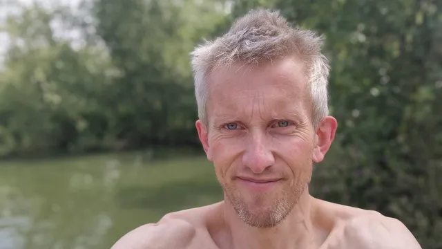 A grey-haired man with a faint smile takes a selfie beside a tree-lined river.