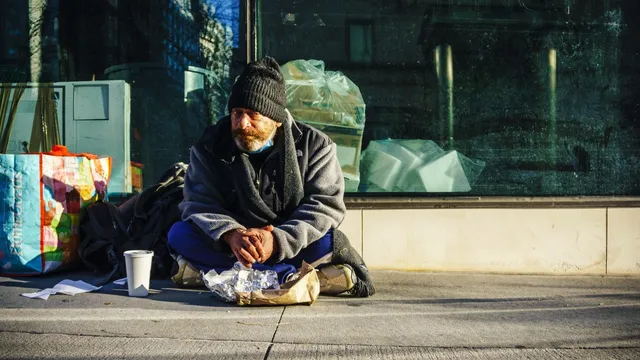 A man in a woolly hat sits cross-legged on a city pavement by a shop window, with bags and a paper cup beside him.
