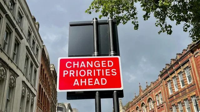 A red road sign reading 'Changed priorities ahead' on a street between brick buildings.