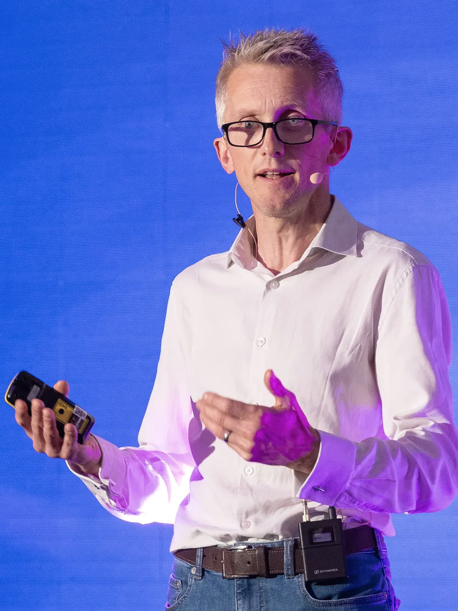 Andrew Pain, in a white shirt, speaking on a stage in front of a blue screen